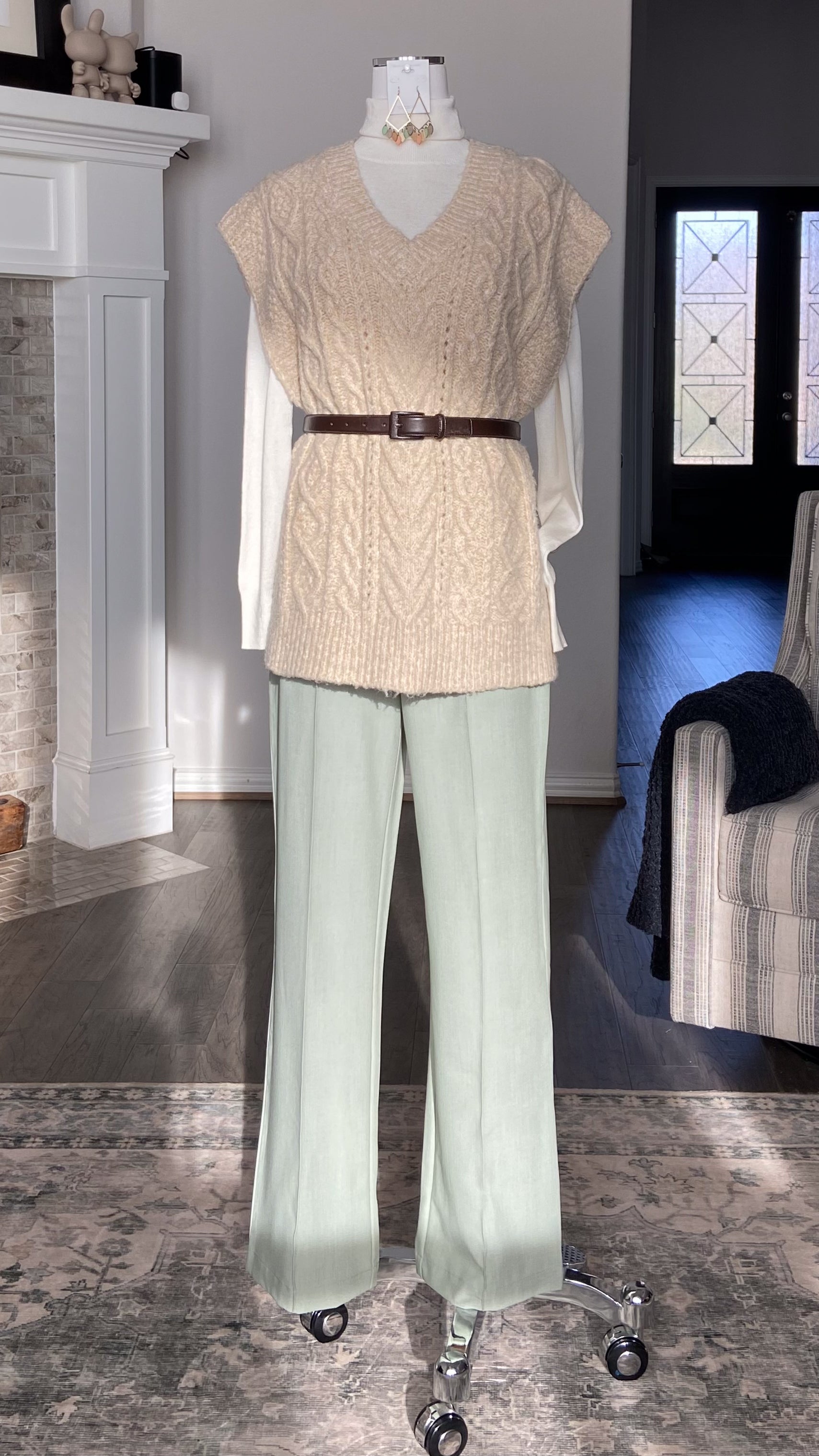 Mannequin wearing a beige knitted top and light green pants in a room with a fireplace and a window.