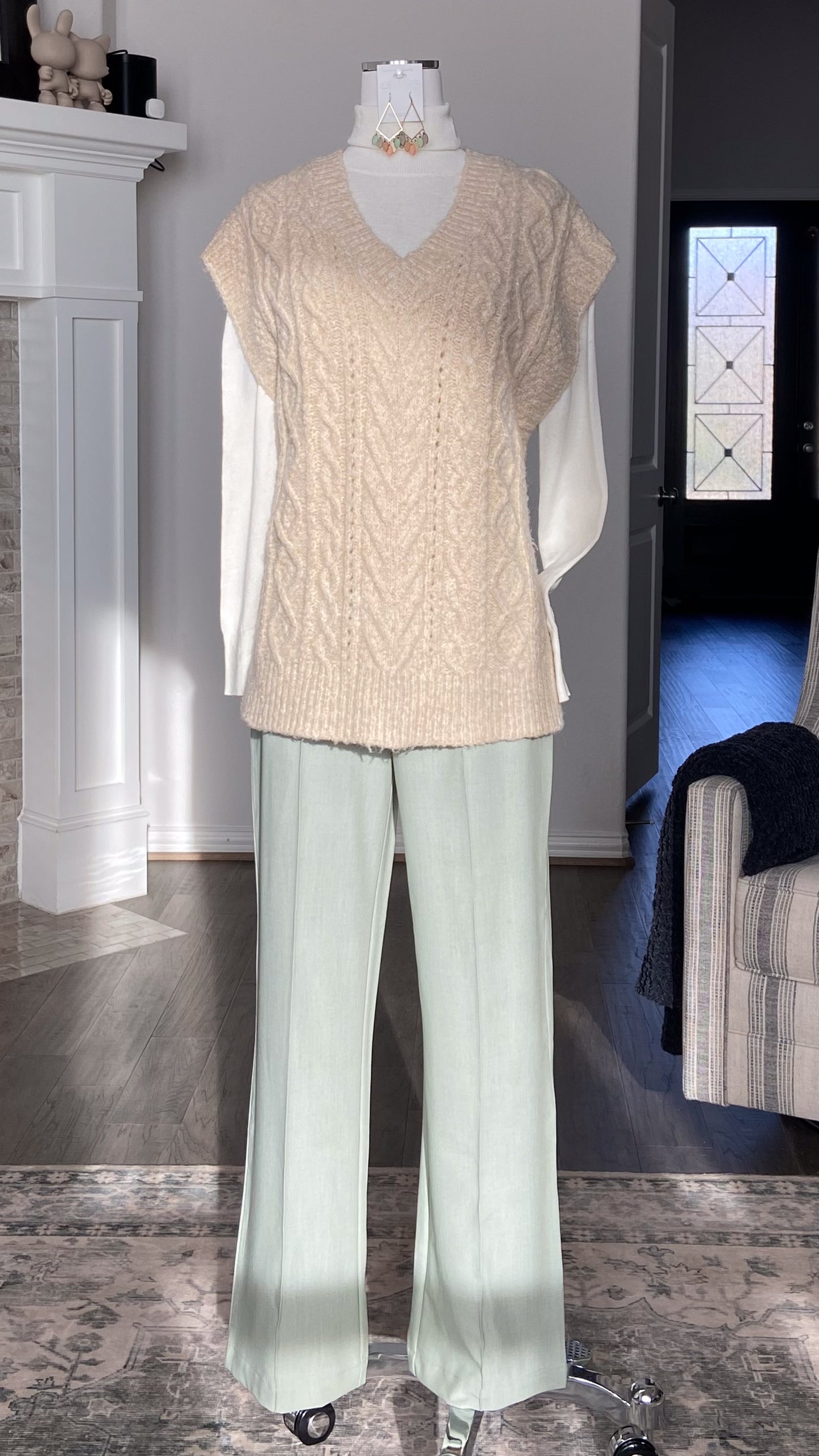 Mannequin wearing a beige V-neck knitted sweater vest and light green pants in a room setting.