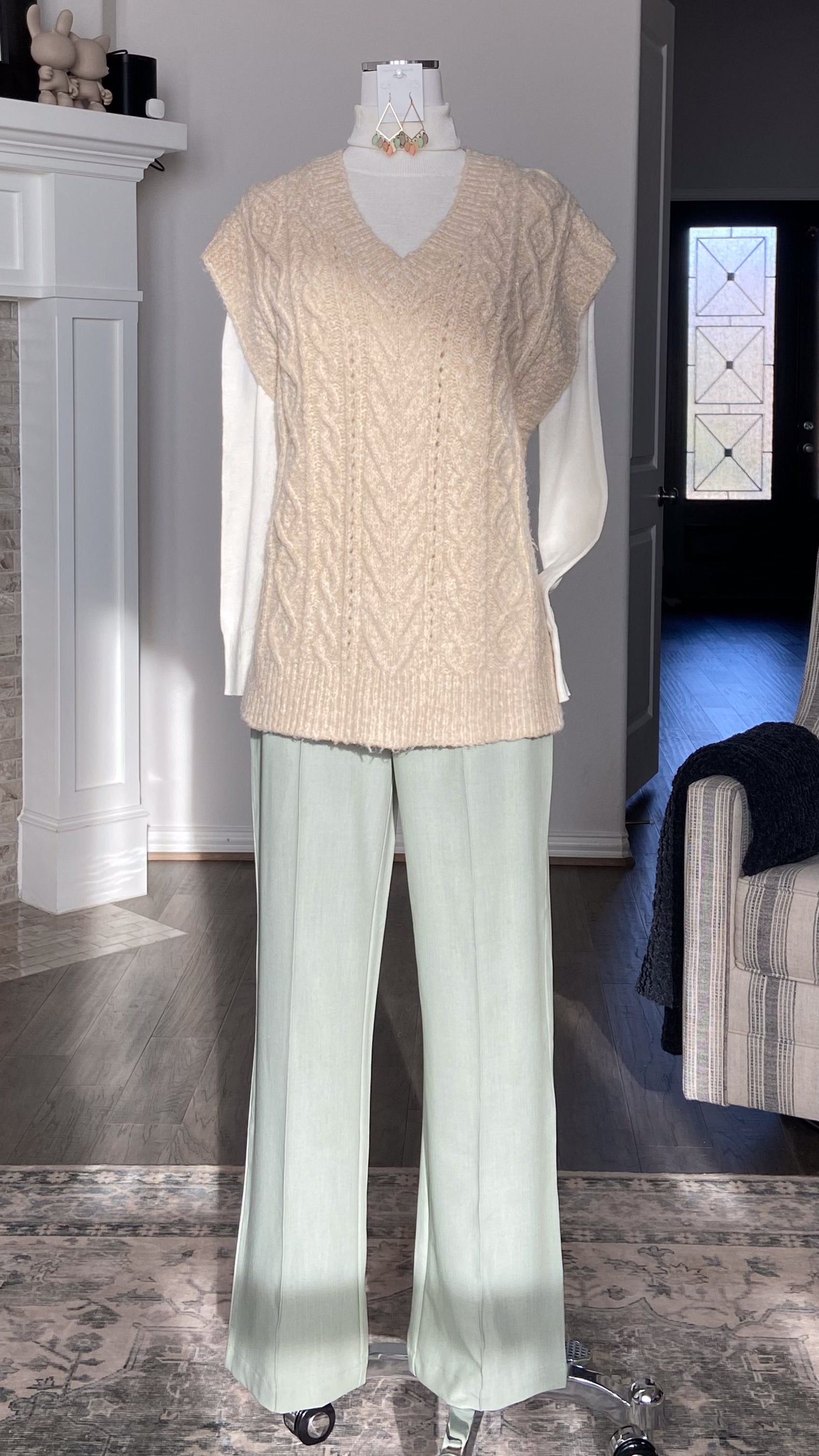 Mannequin wearing a beige V-neck knitted sweater vest and light green pants in a room setting.